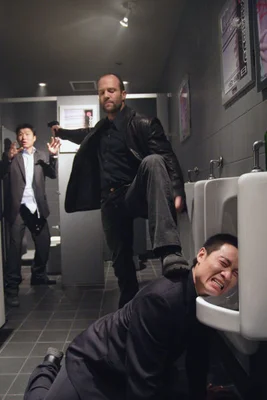 Jason Statham en veste noire en cuir donnant un coup de pied violent sur la tête d'un homme vers un urinoir dans une scène d'action du film Rogue (War).