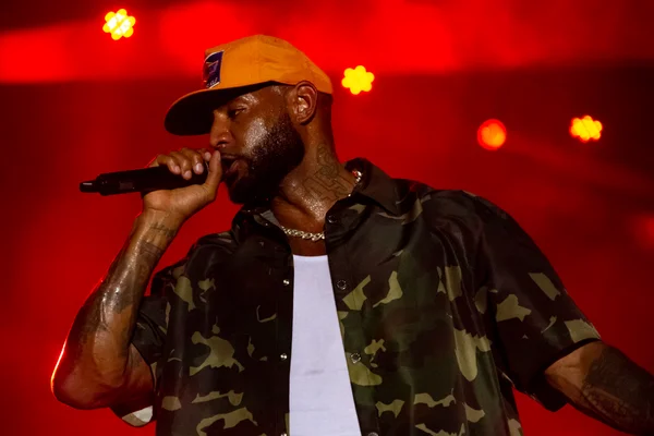 Le rappeur Booba en prestation sur la scène du FEMUA 15 le 29 avril 2023, portant une casquette orange et une chemise camouflage, chantant dans un micro sous un éclairage rouge