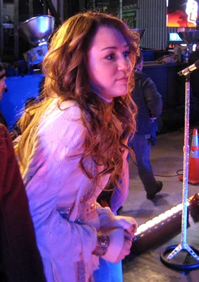 Miley Cyrus, cheveux longs ondulés châtain roux, portant un pull blanc texturé, près d'un micro dans une salle mal éclairée aux lumières bleues et violettes, essayant de se réchauffer avant l'enregistrement de la tournée Hannah Montana Best of Both Worlds (2007/08).