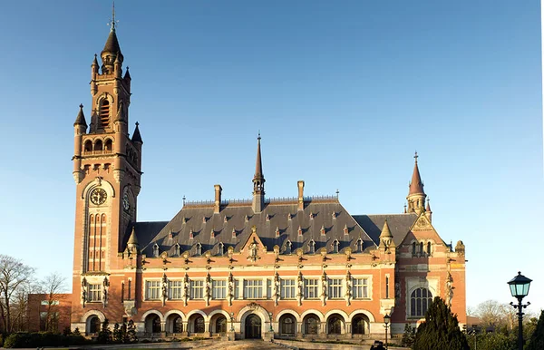 Grand bâtiment de briques rouges de style néo-Renaissance avec tour horloge et statues sous ciel bleu, siège de la Cour internationale de justice à La Haye.