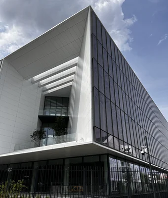 Le nouveau bâtiment du Centre International de Recherche sur le Cancer (CIRC/IARC) à Gerland, Lyon, structure moderne de plusieurs étages avec façade blanche épurée, ouverture rectangulaire en retrait et grande verrière, vu de bas sous un ciel bleu nuageux.