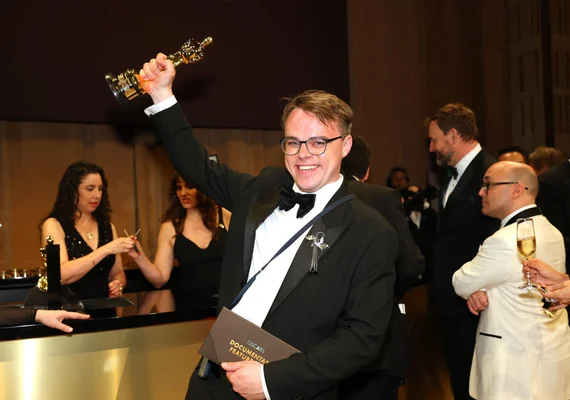Le réalisateur du film "Mr. Nobody Against Putin" en smoking et lunettes, souriant et levant une statuette Oscar lors d'une cérémonie de remise de prix, avec des invités en arrière-plan.