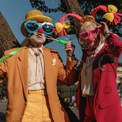 Guide d'idées de soirée déguisée du blog Bamboula : deux personnes âgées en costumes de clown colorés avec lunettes géantes et nez rouge, soufflant des bulles près d'un arbre.