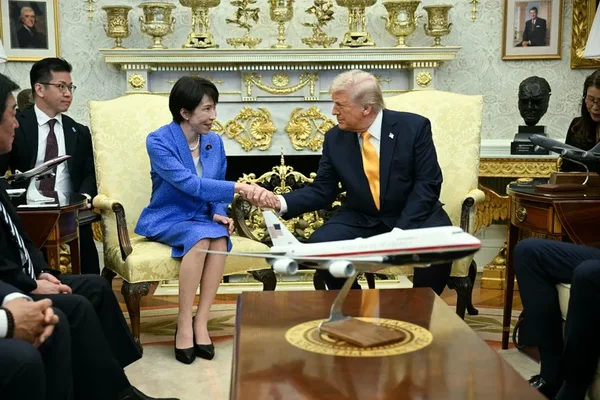 Donald Trump serrant la main de la première ministre japonaise Takaichi en costume bleu, assis dans des fauteuils ornés avec un modèle d'avion sur la table, lors de leur rencontre sur Pearl Harbor.