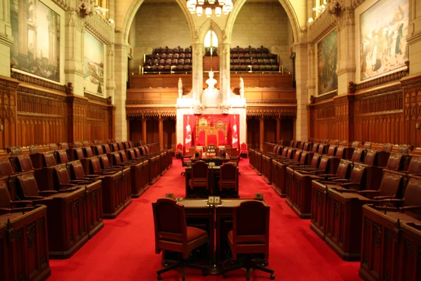 La chambre du Sénat du Canada, présentant des rangées de chaises en cuir rouge, des panneaux de bois sombre et une estrade centrale, salle vide soulignant les détails architecturaux et moquette rouge.