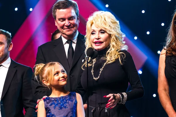 Dolly Parton, en robe noire et collier, parlant au microphone sur scène à l'hôtel Universal Hilton lors des Movieguide Awards, avec Alyvia Alyn Lind en robe bleue la regardant et Stephen Herek près d'eux.