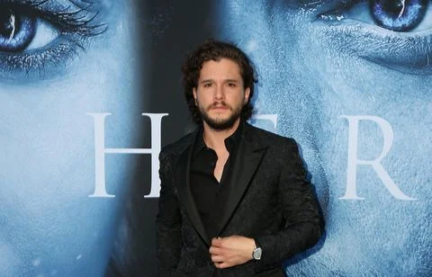 Kit Harington se tenant devant une affiche promotionnelle du film 'Her', vêtu d'une veste à motifs sombres, d'une chemise noire et d'une montre argentée.