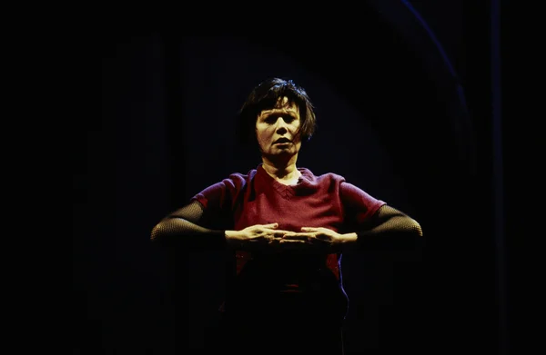 Guesch Patti sur scène lors du spectacle "Elle sourit aux larmes", portant un t-shirt bordeaux et des manches en résille noire, les mains jointes devant la poitrine.