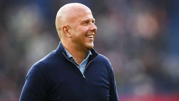 Arne Slot, homme chauve souriant en pull à fermeture éclair marine sur chemise bleu clair, regardant à droite avec une foule floue en arrière-plan, à son arrivée comme entraîneur de Liverpool.