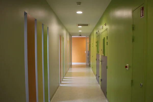 Couloir vide d'une unité pénitentiaire spécialisée en psychiatrie, avec des murs verts à droite et des bandes verticales colorées à gauche menant à un mur orange.
