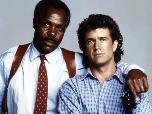 Portrait studio de Danny Glover et Mel Gibson, acteurs du film Lethal Weapon, posant ensemble sur fond gris lors d'une séance photo.