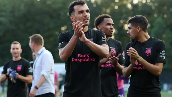 Groupe de joueurs du Sabah FK, jeune club azéri qualifié pour la première fois pour la phase de ligue de la Ligue des champions, en tenues d'entraînement noires sponsorisées par Bank Respublika, applaudissant sur un terrain herbeux avec des arbres.