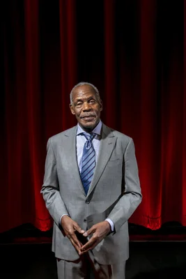 Danny Glover en costume gris avec une cravate rayée bleue, les mains jointes devant des rideaux de théâtre rouges.