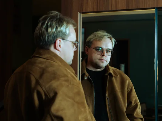 Homme aux cheveux blonds et lunettes, portant une veste en daim marron sur chemise noire, se regardant dans un miroir, en lien avec le documentaire oscarisé "Mr Nobody contre Poutine" visé par les autorités russes.