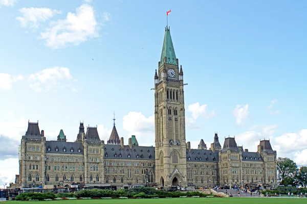 La Colline du Parlement à Ottawa, sur les berges du fleuve Ottawa et siège du Parlement du Canada, montrant le bloc central néogothique avec la tour de la Paix et le drapeau canadien sous un ciel bleu, pelouse verte à l'avant.