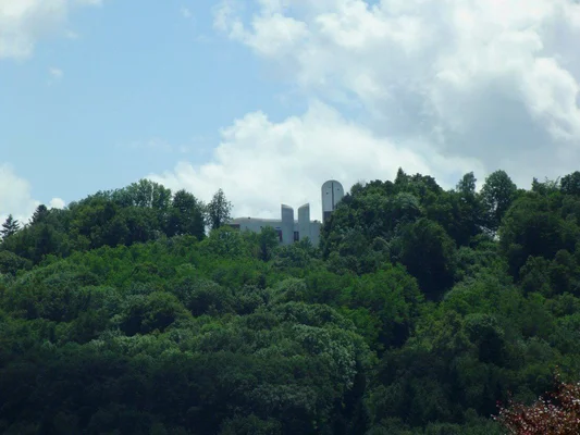 La chapelle Le Corbusier située sur la colline de Bourlémont à Ronchamp, bâtiment moderne blanc à tour cylindrique et ailettes verticales, visible sur une crête partiellement masquée par de denses arbres verts sous un ciel bleu nuageux.