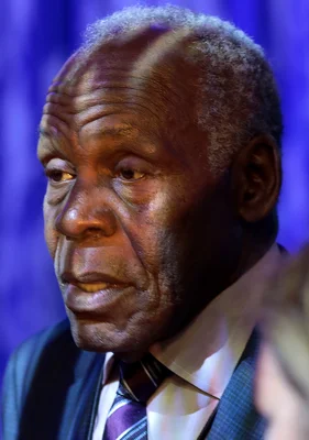 Gros plan de l'acteur Danny Glover, un homme plus âgé à cheveux gris courts, vêtu d'un costume et d'une cravate, regardant légèrement vers la gauche sur un arrière-plan bleu flou.