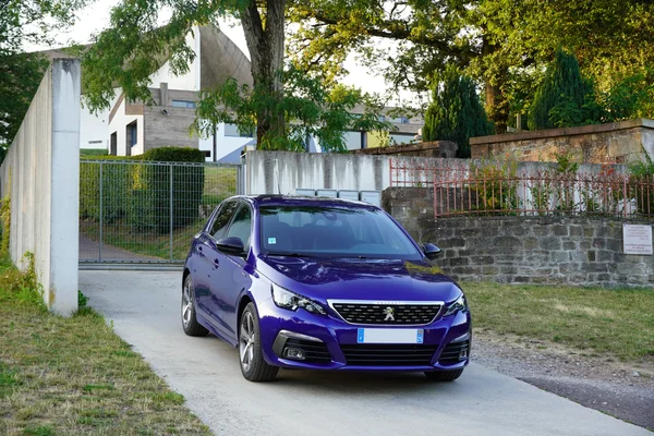 Berline violette Peugeot 308 de 2020 garée sur une voie pavée près d'un mur en pierre et d'une clôture, devant la chapelle Notre-Dame-du-Haut de Ronchamp, bâtiment moderne en arrière-plan.