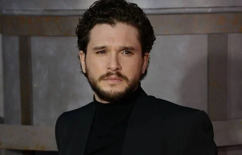 Kit Harington, acteur aux cheveux bouclés et à la barbe, portant un col roulé noir et une veste sur fond flou, dans un portrait tête et épaules.