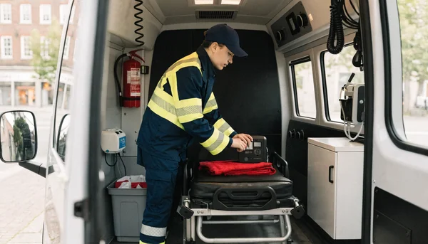 Illustration d'un jeune stagiaire ambulancier en uniforme à l'intérieur d'une ambulance, concentré sur la vérification d'un moniteur médical posé sur un brancard, éclairé par la lumière du jour.