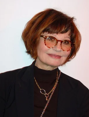 Portrait récent de Guesch Patti avec des cheveux auburn courts, portant un blazer noir, un col roulé, des colliers et des lunettes à monture écaille avec des verres teintés en rouge.