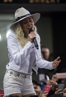 Miley Cyrus, aux cheveux blonds, portant un chapeau blanc, une veste en jean blanche et un short blanc, chantant dans un microphone sur scène lors de l'émission Today Show à Manhattan pour la Fleet Week 2017.