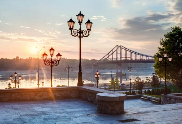 Passerelle piétonne de Parkovyi à Kiev, en Ukraine, montrant une rivière au lever ou coucher du soleil, un grand pont suspendu en arrière-plan et des lampadaires ornés au premier plan.