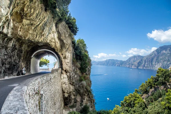 Route côtière pittoresque traversant un tunnel taillé dans une falaise escarpée, surplombant une mer bleue vif et des montagnes lointaines sous un ciel clair.