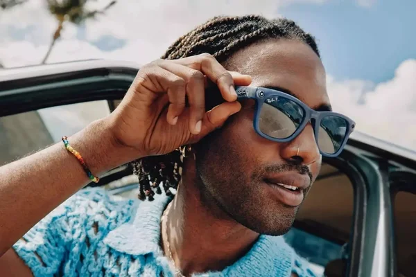 Un homme aux tresses portant une chemise bleue en crochet ajuste ses lunettes de soleil bleues Ray-Ban Meta tout en se penchant par la fenêtre d'une voiture.