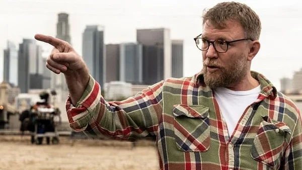 Guy Ritchie portant une chemise à carreaux et des lunettes, pointe du doigt vers la gauche devant un panorama urbain.