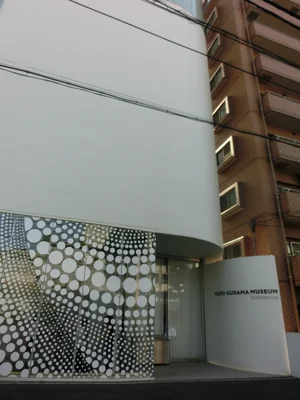 Façade extérieure du Yayoi Kusama Museum à Tokyo, avec sa structure courbe blanche et une entrée en verre décorée de points blancs polka-dot.
