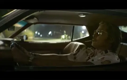 Un homme portant des lunettes de soleil et une chemise à motifs conduisant une voiture de nuit, avec une expression détendue.
