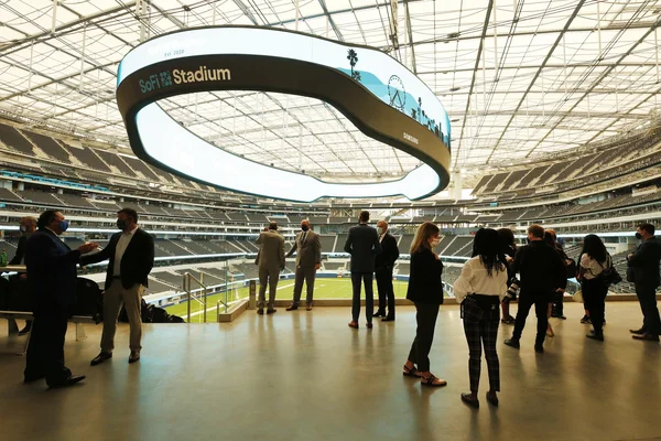 Un groupe de personnes en tenue d'affaires se tient sur un balcon donnant sur l'intérieur du SoFi Stadium, admirant l'écran vidéo circulaire suspendu.