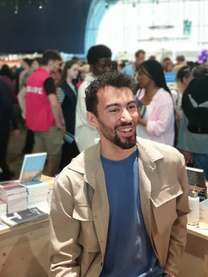 Nassim Kezoui, auteur de "Agrégat. Face A", souriant à un événement littéraire, assis à une table entourée de livres, vêtu d'une veste beige sur un t-shirt bleu