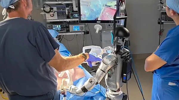 Un chirurgien en blouse bleue opérant un système chirurgical robotique tandis qu'un autre professionnel observe, avec un moniteur affichant une diffusion en direct du site chirurgical.