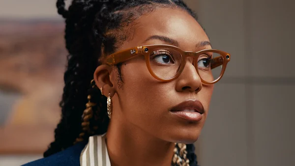 Une jeune femme aux tresses coiffée de lunettes Ray-Ban Meta ambrées, portant des boucles d'oreilles en or et un blazer bleu sur une chemise rayée, regardant vers le côté.