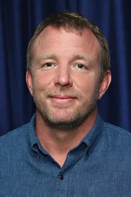 Portrait de Guy Ritchie, homme d'affaires aux cheveux bruns courts et à la barbe légère, en chemise bleue boutonnée devant un rideau foncé.
