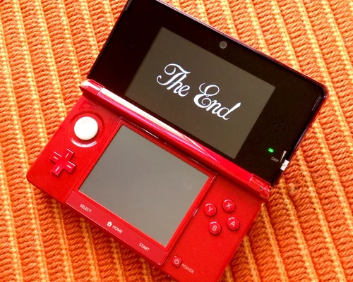 Un Nintendo 3DS XL rouge ouvert sur une surface orange, affichant le texte « The End » en police blanche et cursive sur son écran supérieur, montrant la fin du jeu Dragon Quest VII pour la Nintendo 3DS.