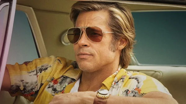 Brad Pitt dans le rôle de Cliff Booth, assis au volant d'une voiture, portant une chemise florale jaune, des lunettes de soleil et une montre dorée, lors de l'annonce du titre du film sur Netflix.