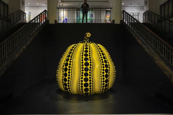 Sculpture de citrouille jaune géante à pois noirs de Yayoi Kusama exposée dans une salle sombre et symétrique avec des escaliers.