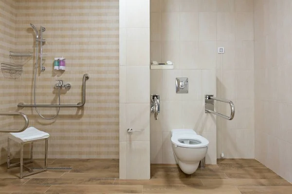 Salle de bain aménagée pour personnes handicapées avec douche de plain-pied équipée d'une assise rabattable et barres d'appui à gauche, et WC suspendu avec barres de soutien à droite, carrelage beige et sol effet bois