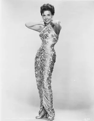 Portrait en studio en noir et blanc de Lena Horne, circa 1950, portant une robe à bretelles croisées à motif, posant avec les mains derrière la tête.