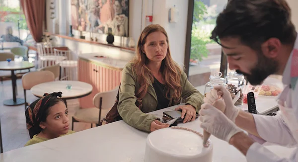 Scène du film égyptien "Happy Birthday" montrant une femme et une jeune fille observant attentivement un pâtissier décorer un gâteau blanc au comptoir d'un café