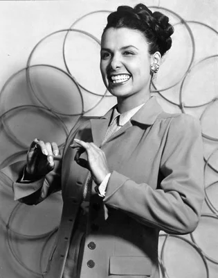 Photographie en noir et blanc de Lena Horne, circa 1940, avec des cheveux sombres coiffés en chignon, portant une veste à col et tenant une ceinture, debout devant un fond de cerceaux circulaires superposés.