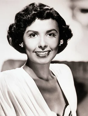 Portrait en studio en noir et blanc de Lena Horne, chanteuse, avec des cheveux courts, bouclés et sombres, portant un haut clair et des boucles d'oreilles, souriant face à la caméra.