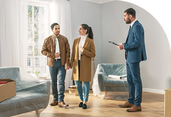 Un agent immobilier en costume bleu, avec un classeur, discute avec un couple souriant dans une pièce lumineuse et vide, dont les meubles sont recouverts de plastique, illustrant la visite d'un bien neuf.