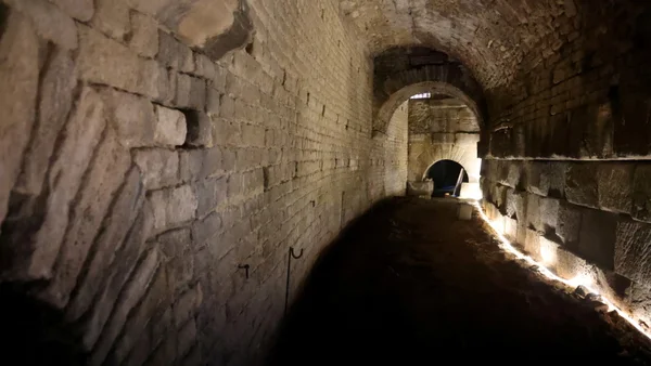 Couloir souterrain voûté des Arènes d'Arles aux murs en pierre brute, faiblement éclairé, s'étendant en perspective vers une arche lumineuse au fond du tunnel sombre