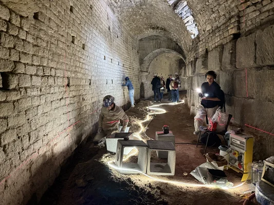 Groupe de techniciens travaillant dans le long tunnel voûté en pierre des souterrains des Arènes d'Arles, utilisant des équipements et une bande lumineuse pour relever et documenter la structure