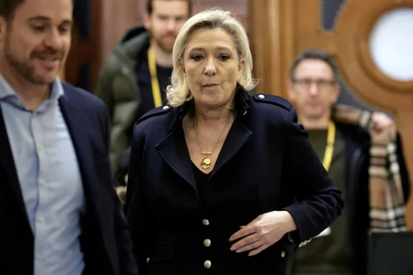 Marine Le Pen vêtue d'une veste bleu marine foncé marchant lors de son procès du Rassemblement national, accompagnée d'un homme en costume à sa gauche et d'autres personnes en arrière-plan
