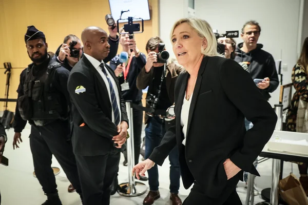 Marine Le Pen, femme blonde en tailleur noir, traversant une salle d'audience entourée de photographes et d'agents de sécurité lors du procès du RN, sous les flashes des appareils photo et les microphones tendus